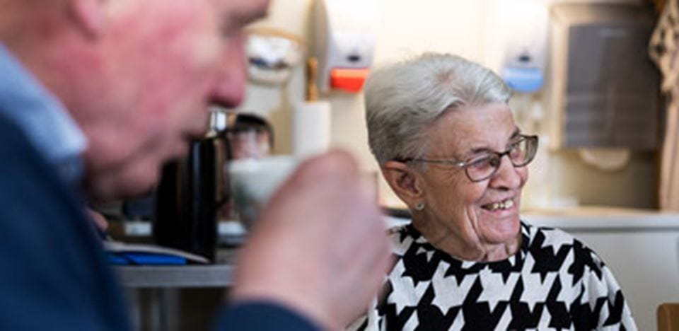 Ældre mand i forgrunden drikker kaffe, ældre dame i baggrunden smiler